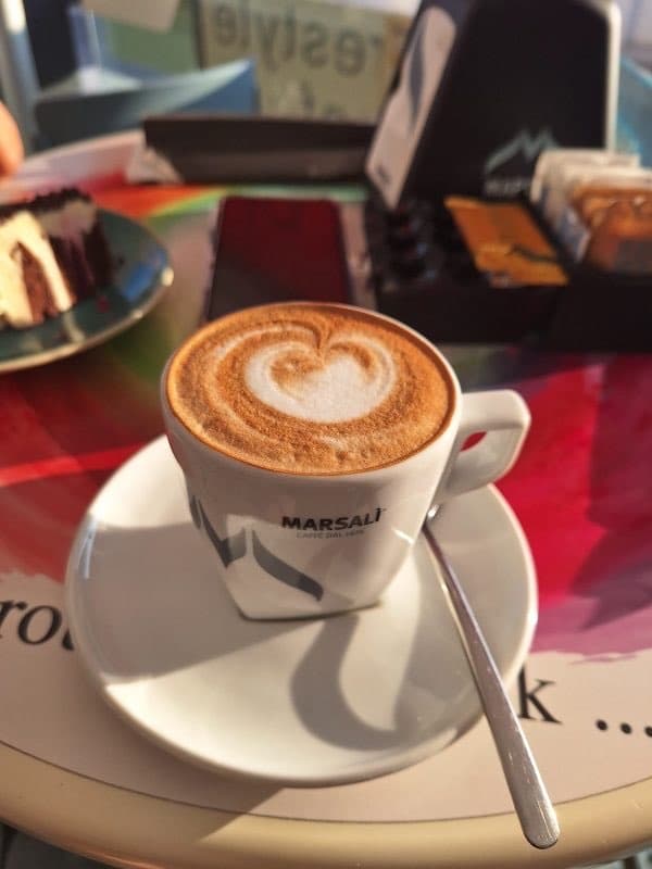 A Marsali cappuccino with a heart on top, on a printed terrace table, casting a long late-afternoon shadow